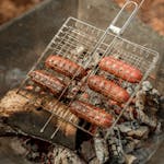 Sizzling sausages being grilled over a smoky outdoor fire for a barbecue.