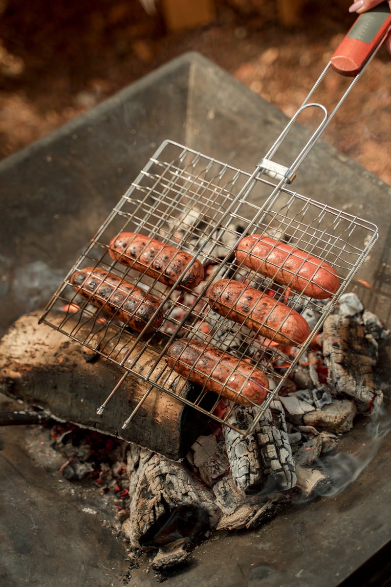 Sizzling sausages being grilled over a smoky outdoor fire for a barbecue.